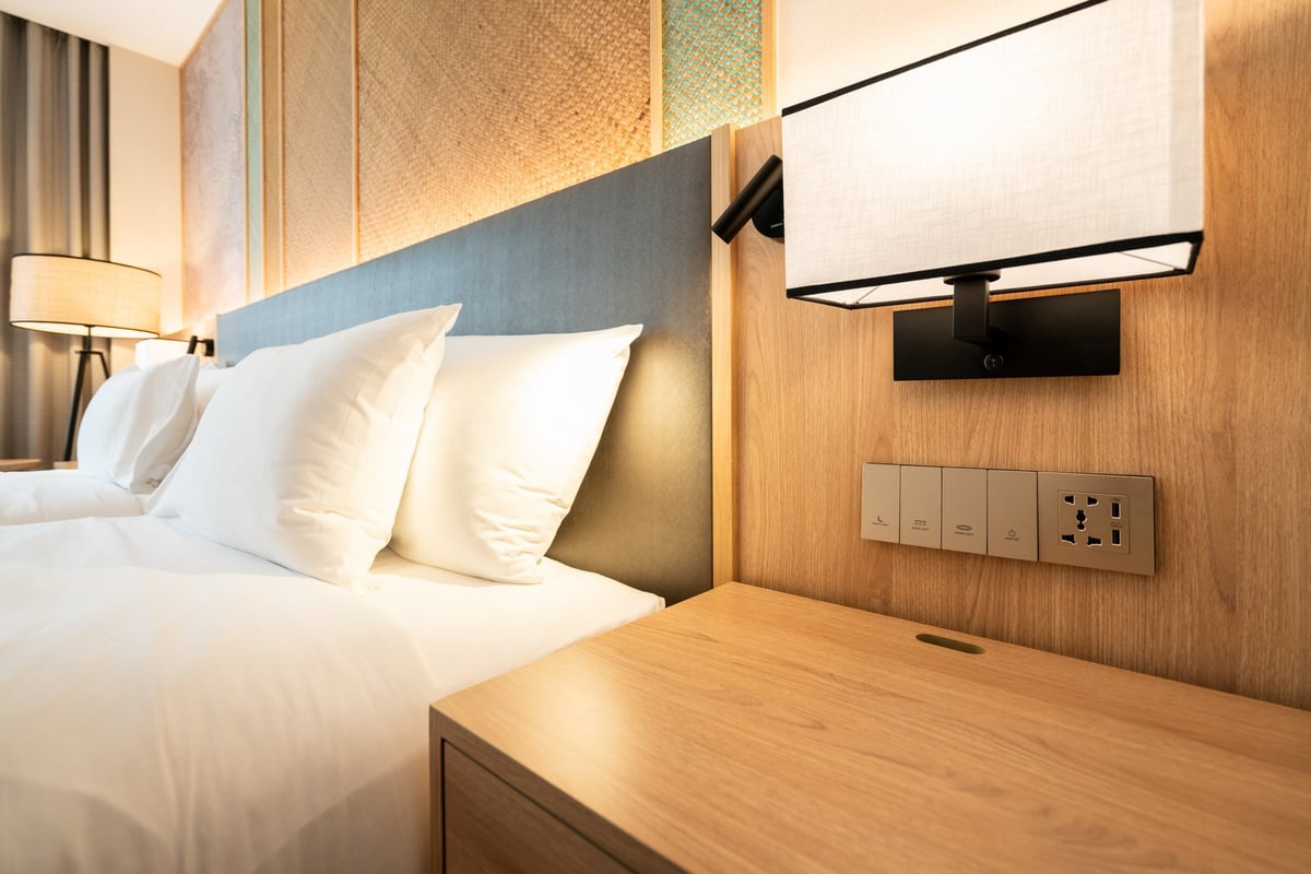 Detail of a clean and modern hotel room showing the bed with white pillows, wooden nightstand, fabric wall lamp, and a power outlet, switch panel.
