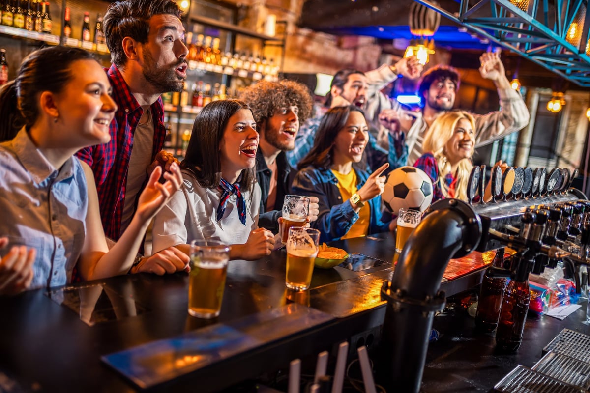 Happy group of fans watching a football game and celebrating a goal in a pub. People with beer glasses and a soccer ball in an urban bar.